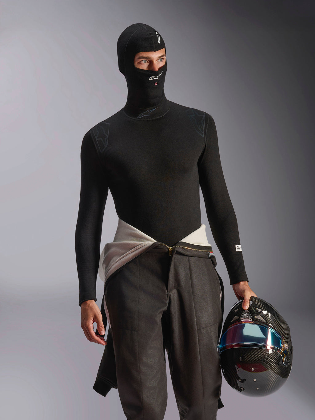 Modelo masculino a usar a Balaclava Alpinestars ZX Evo V3 em preto, com logótipos brancos na testa e no queixo, combinada com uma camisola interior de manga comprida preta e a segurar um capacete de corrida em fibra de carbono, equipamento de automobilismo profissional resistente às chamas.
