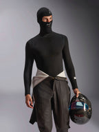 Modelo masculino a usar a Balaclava Alpinestars ZX Evo V3 em preto, com logótipos brancos na testa e no queixo, combinada com uma camisola interior de manga comprida preta e a segurar um capacete de corrida em fibra de carbono, equipamento de automobilismo profissional resistente às chamas.