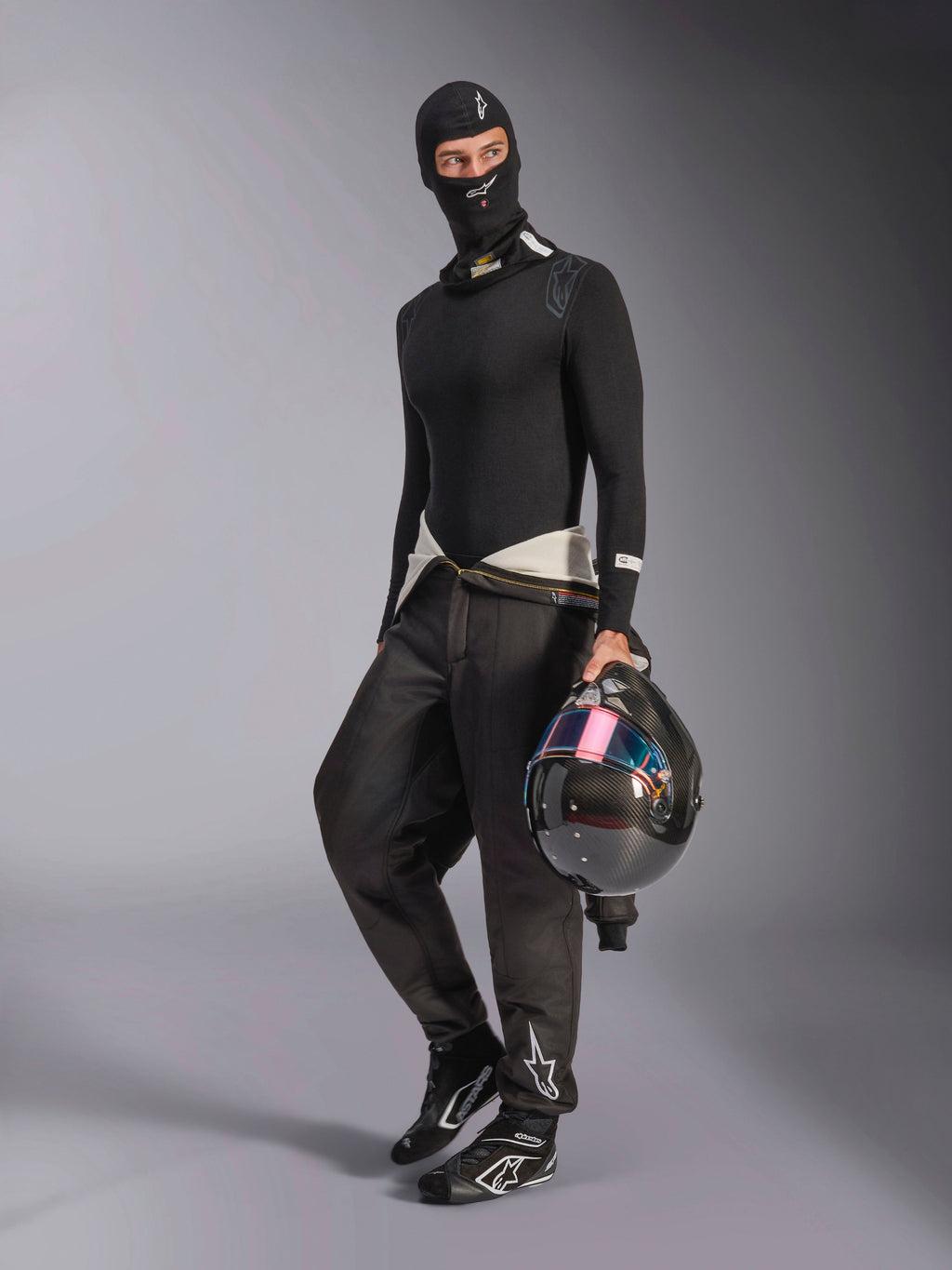Balaclava Alpinestars ZX Evo V3, preta, usada por um piloto com equipamento completo de base layer para automobilismo, com o logótipo branco na testa, a segurar um capacete de fibra de carbono com viseira iridescente, vestuário profissional de desporto motorizado resistente ao fogo.