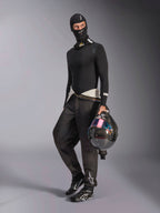 Balaclava Alpinestars ZX Evo V3, preta, usada por um piloto com equipamento completo de base layer para automobilismo, com o logótipo branco na testa, a segurar um capacete de fibra de carbono com viseira iridescente, vestuário profissional de desporto motorizado resistente ao fogo.