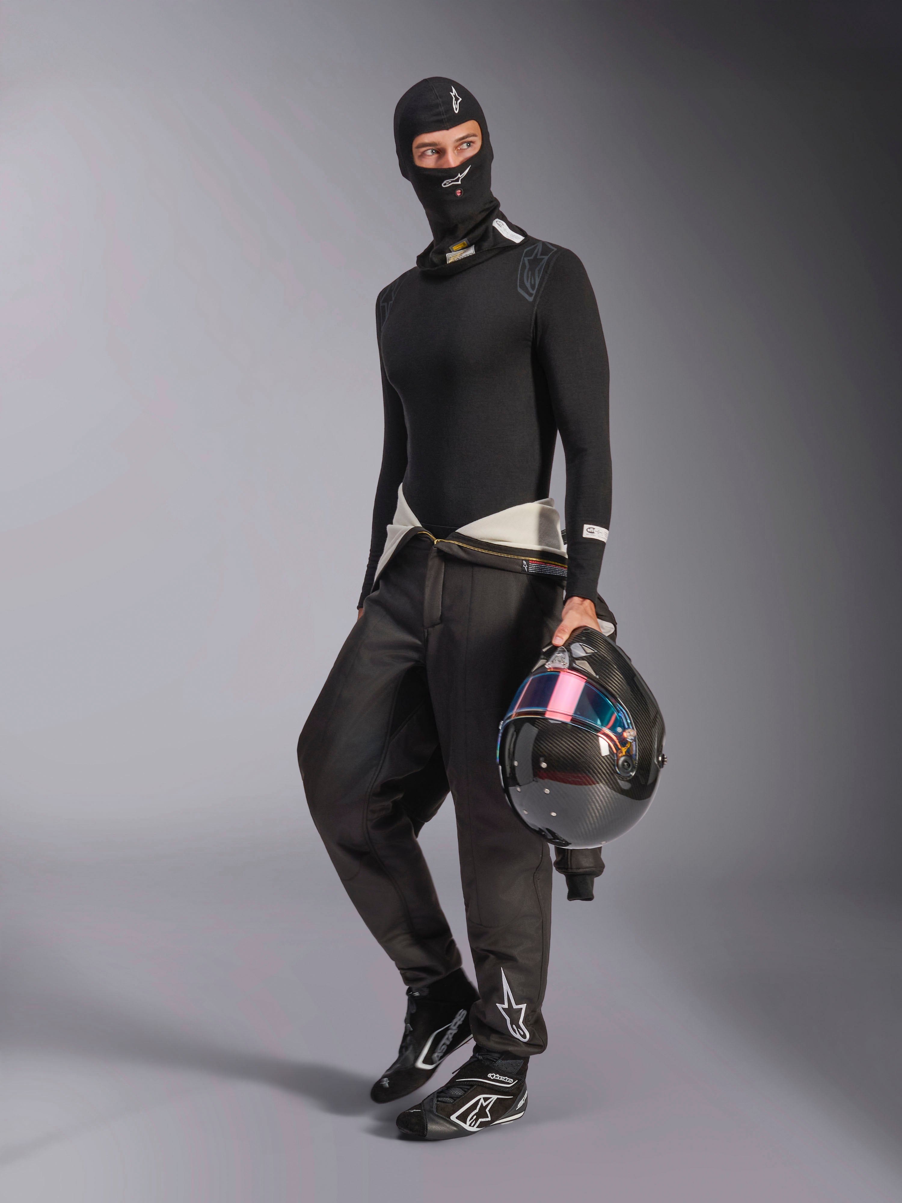 Balaclava Alpinestars ZX Evo V3, preta, usada por um piloto com equipamento completo de base layer para automobilismo, com o logótipo branco na testa, a segurar um capacete de fibra de carbono com viseira iridescente, vestuário profissional de desporto motorizado resistente ao fogo.