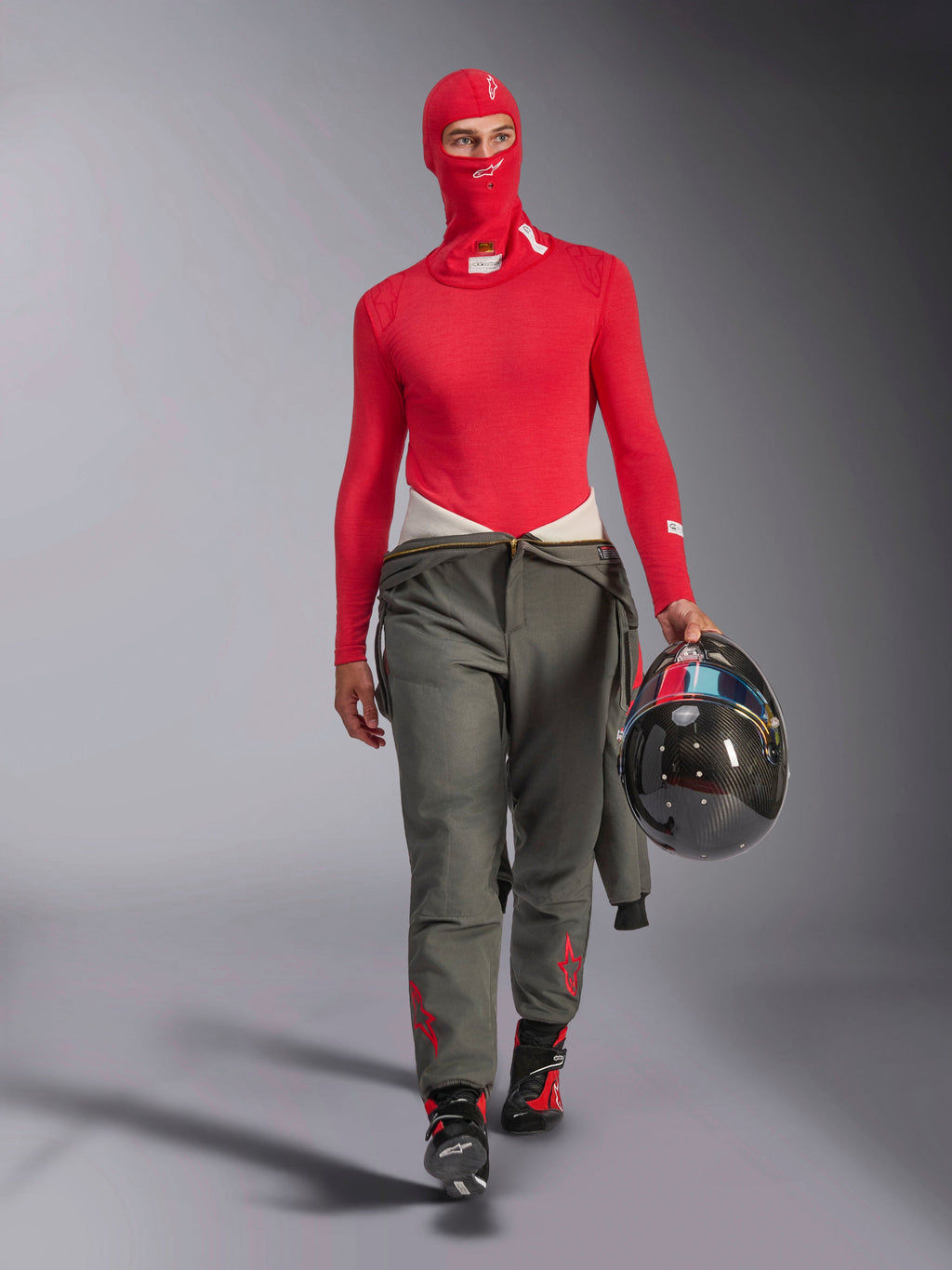 Alpinestars ZX Evo V3 Balaclava em Vermelho, usada por um piloto de corridas com uma camada de base de manga comprida vermelha a condizer e calças de corrida cinzentas, segurando um capacete de fibra de carbono, equipamento de automobilismo profissional resistente às chamas