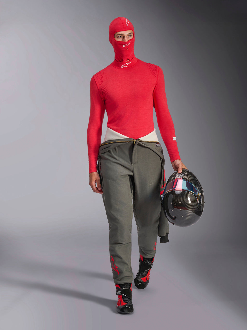 Piloto de corridas a usar balaclava Alpinestars ZX Evo V3 em vermelho, camada de base de manga comprida vermelha a condizer, calças de corrida cinzentas, botas de corrida pretas e vermelhas, a segurar um capacete de fibra de carbono, conjunto de equipamento profissional de automobilismo