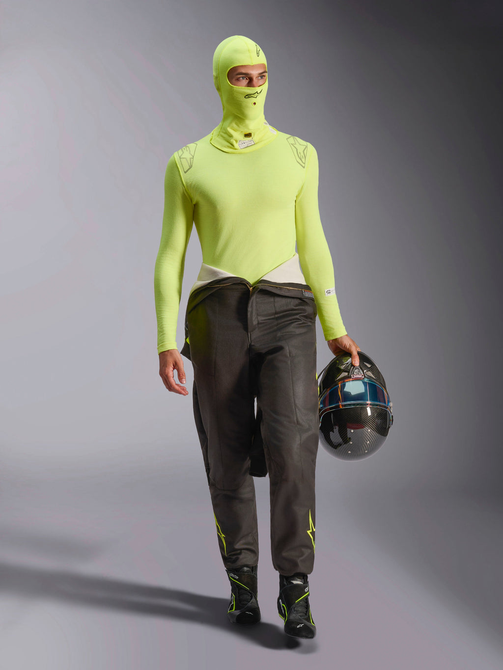 Piloto de corridas masculino a usar a Balaclava Alpinestars ZX Evo V3 em Yellow Fluo, camisola de base de manga comprida a condizer, calças de corrida escuras e botas de corrida pretas, a segurar um capacete de fibra de carbono, equipamento profissional de automobilismo