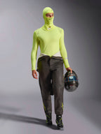 Piloto de corridas masculino a usar a Balaclava Alpinestars ZX Evo V3 em Yellow Fluo, camisola de base de manga comprida a condizer, calças de corrida escuras e botas de corrida pretas, a segurar um capacete de fibra de carbono, equipamento profissional de automobilismo