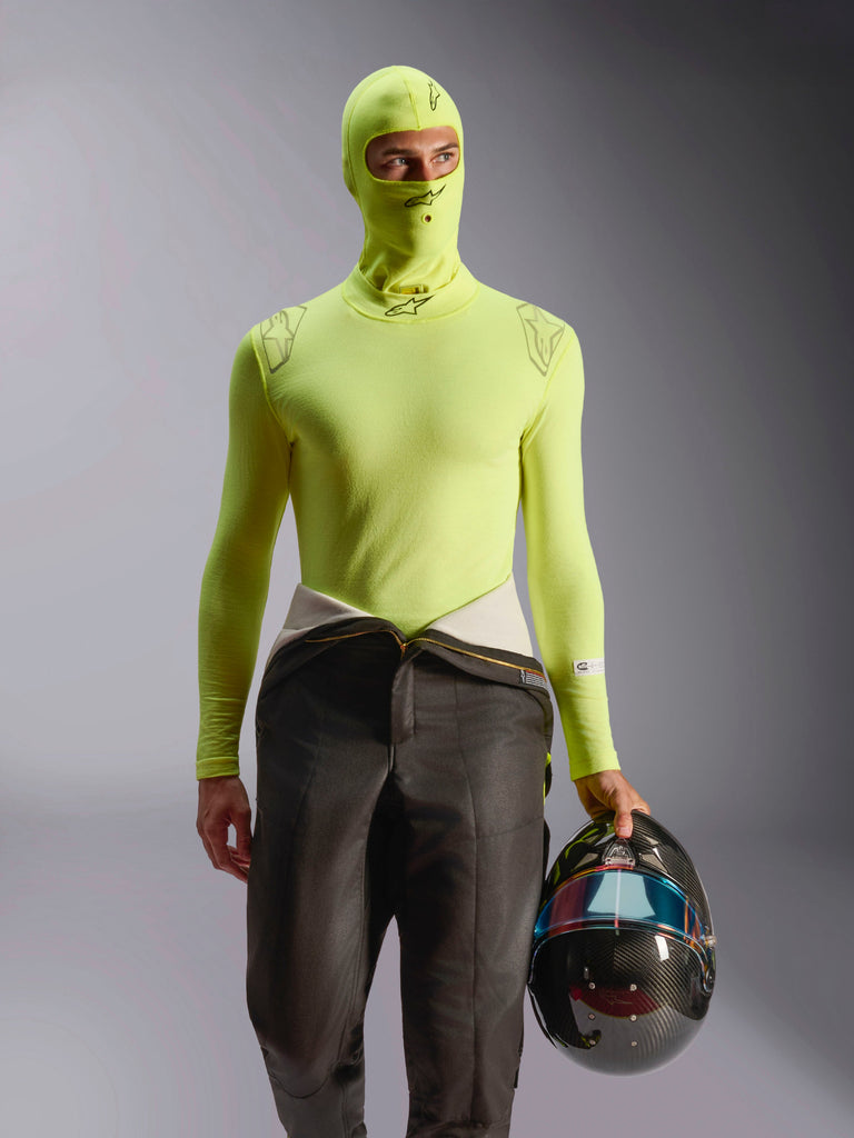 Alpinestars ZX Evo V3 Balaclava, camada de base para automobilismo, amarelo fluo, usada com camisola de manga comprida e calças de fato de corrida a condizer, modelo a segurar um capacete de fibra de carbono com viseira iridescente, equipamento profissional de desporto motorizado resistente ao fogo
