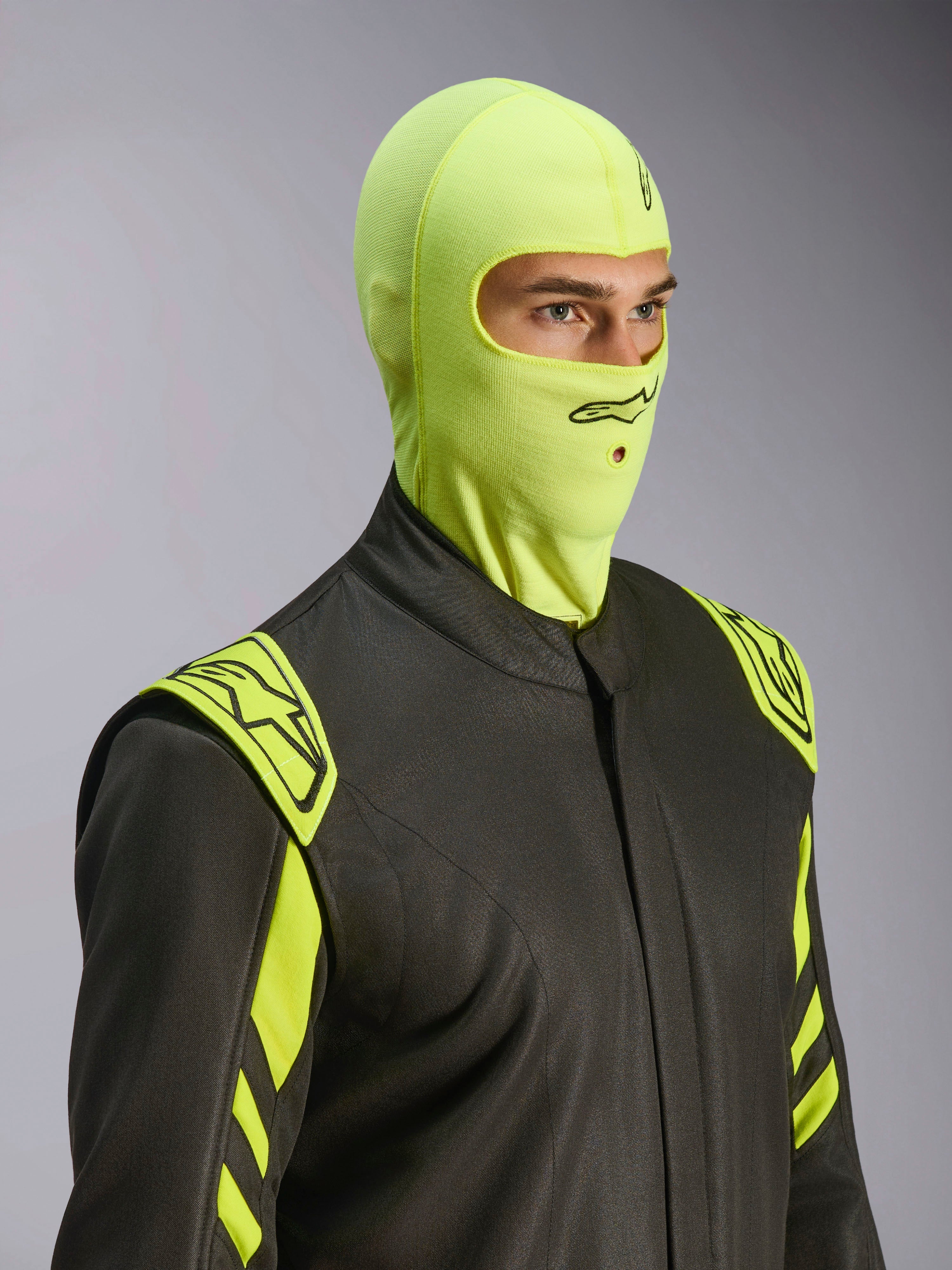 Modelo masculino a usar a balaclava Alpinestars ZX Evo V3 em Yellow Fluo, proteção de cabeça resistente ao fogo para competição, combinada com fato de automobilismo cinzento escuro com detalhes em amarelo néon, logótipo Alpinestars preto e abertura para tubo de hidratação, equipamento de proteção profissional para desportos motorizados