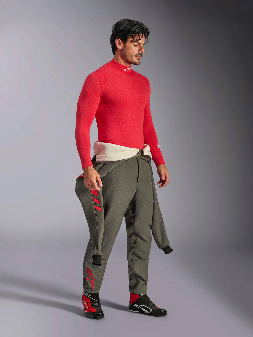 Alpinestars ZX Evo V3 Top de Manga Comprida em Vermelho, camada de base resistente ao fogo para automobilismo, usado com calças de corrida verde azeitona e sapatos de corrida pretos e vermelhos, equipamento profissional de desporto motorizado, retrato de estúdio