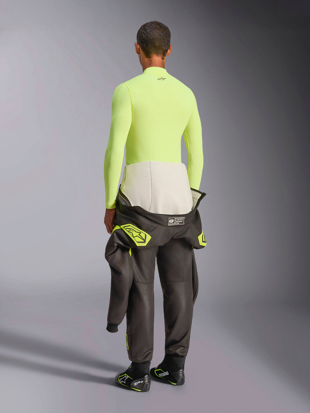 Alpinestars ZX Evo V3 Top de Manga Comprida, Amarelo Fluo, vista traseira de modelo a usar camada de base resistente ao fogo com calças de fato de corrida escuras e botas, roupa interior técnica profissional para desportos motorizados