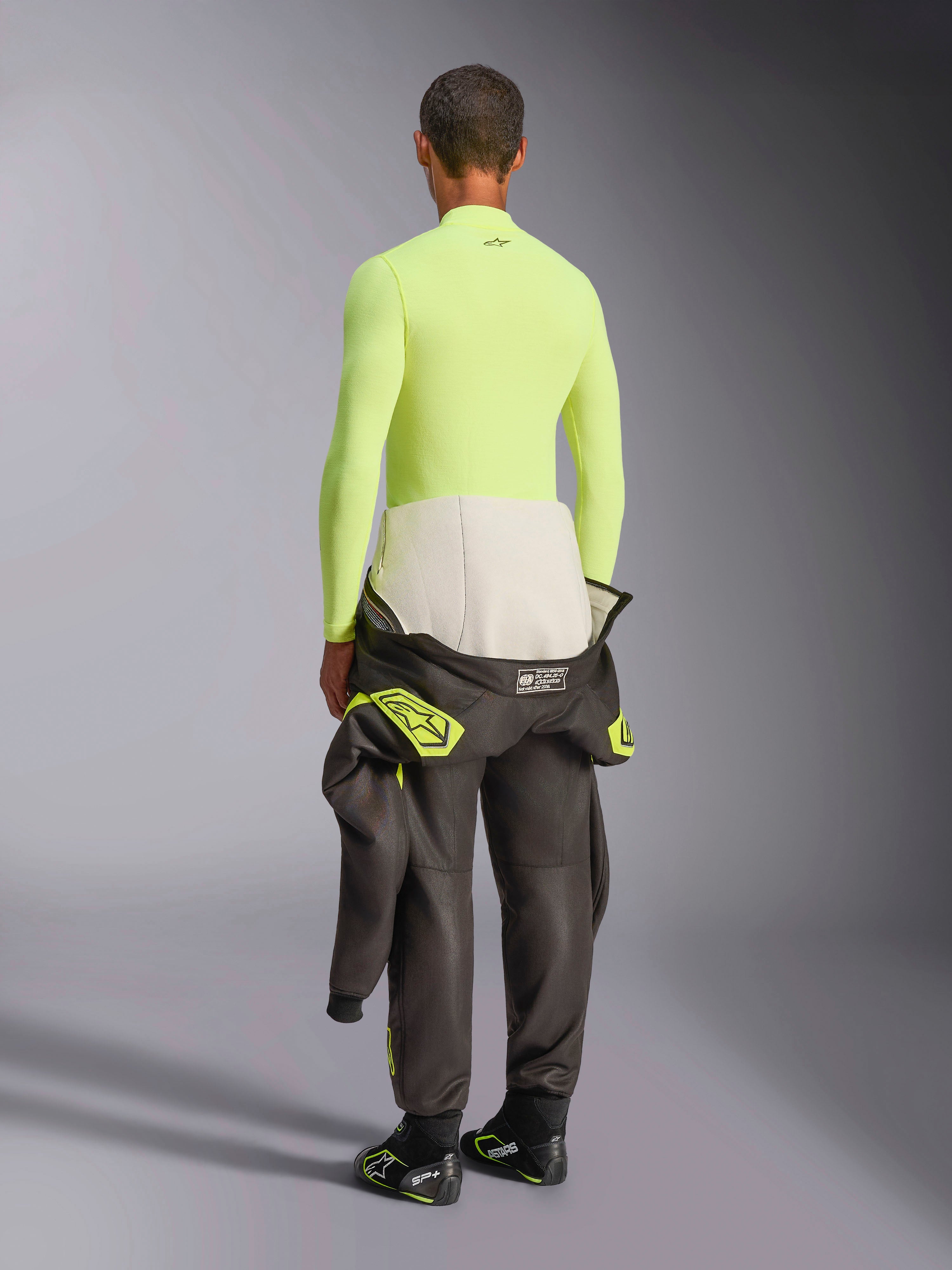 Alpinestars ZX Evo V3 Top de Manga Comprida, Amarelo Fluo, vista traseira de modelo a usar camada de base resistente ao fogo com calças de fato de corrida escuras e botas, roupa interior técnica profissional para desportos motorizados