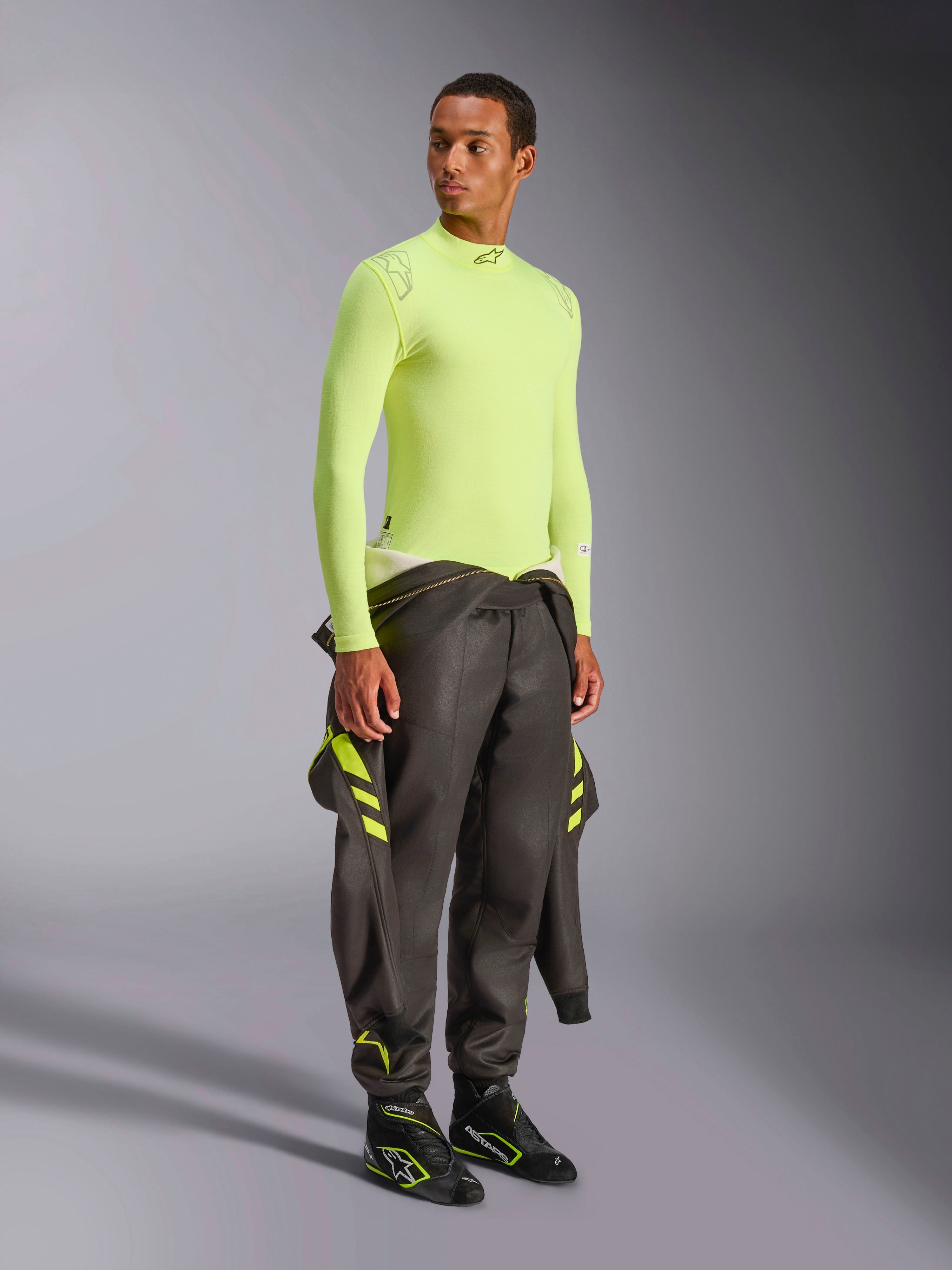Alpinestars ZX Evo V3 Top - Manga Comprida, Amarelo Fluo, vestido por um modelo com um fato de competição cinzento escuro atado à cintura e botas de competição pretas, camada de base de automobilismo resistente às chamas, construção leve em malha jacquard, equipamento profissional de automobilismo