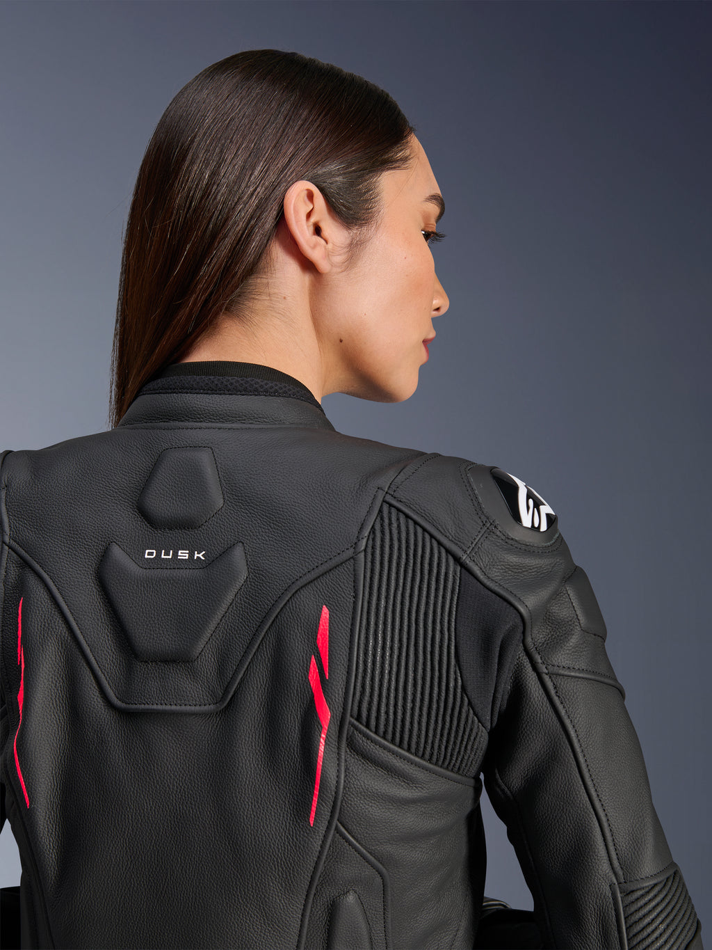 Mulher a usar Casaco de Couro Alpinestars Stella Dusk, couro preto com detalhes Diva Pink e logótipo branco no ombro, vista traseira a mostrar a marca DUSK e painéis elásticos em acordeão, design ergonómico desportivo