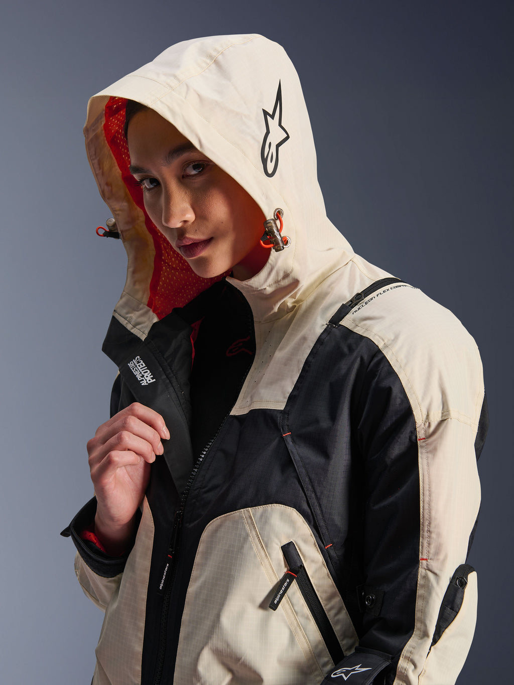 Alpinestars Molly Air Jacket in Black Almond, worn by a female model with the hood up, featuring cream and black color-blocking, a vibrant orange mesh lining, and technical urban design elements including a star logo and zip details.