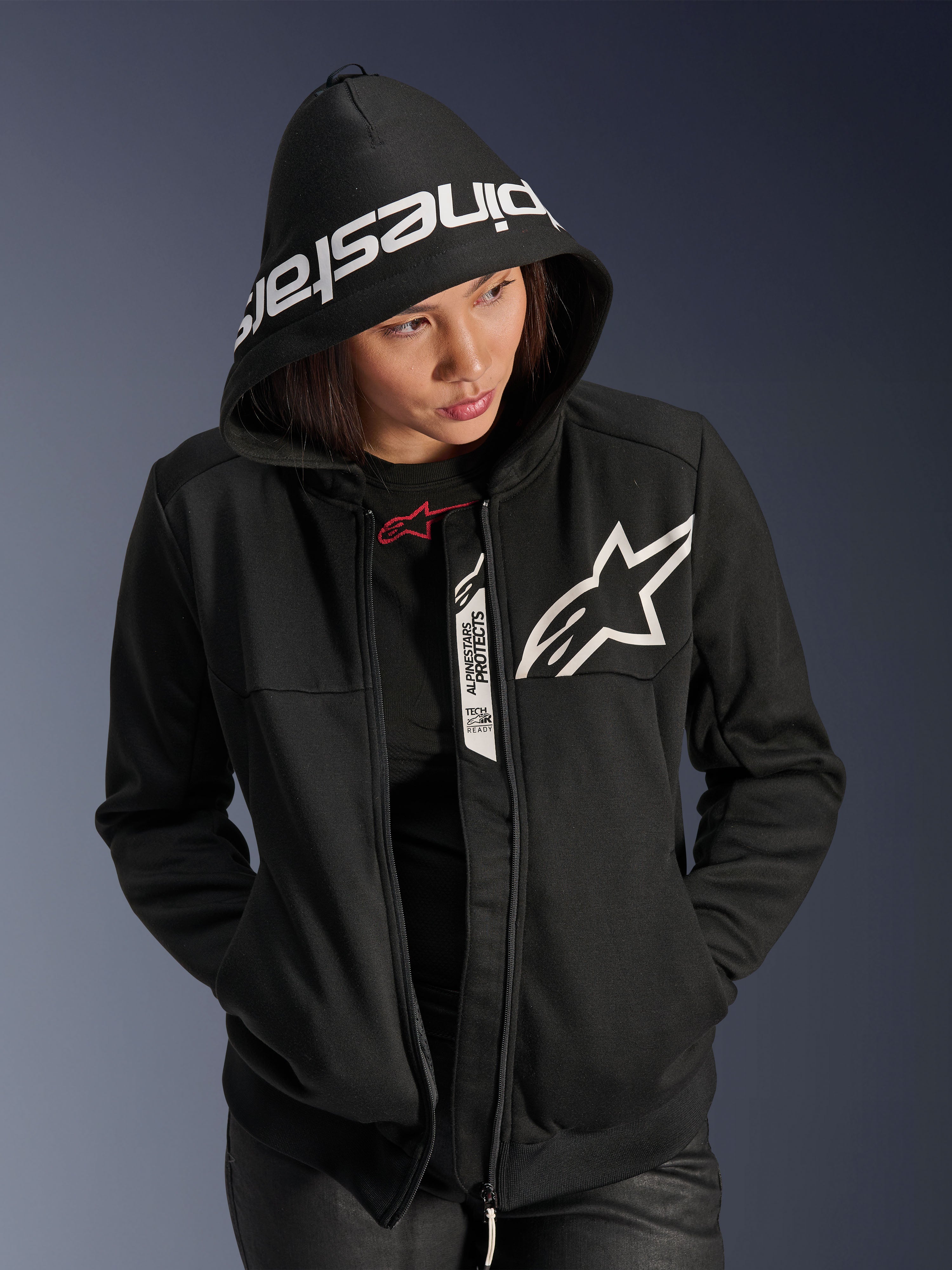 Alpinestars Stella Chrome V2 Sport Hoodie para mulher, preto e branco, hoodie de motociclismo com logótipo de estrela branco no peito e marca Alpinestars na aba do capuz, mulher com o capuz colocado, estilo desportivo urbano casual com detalhes Tech-Air Ready