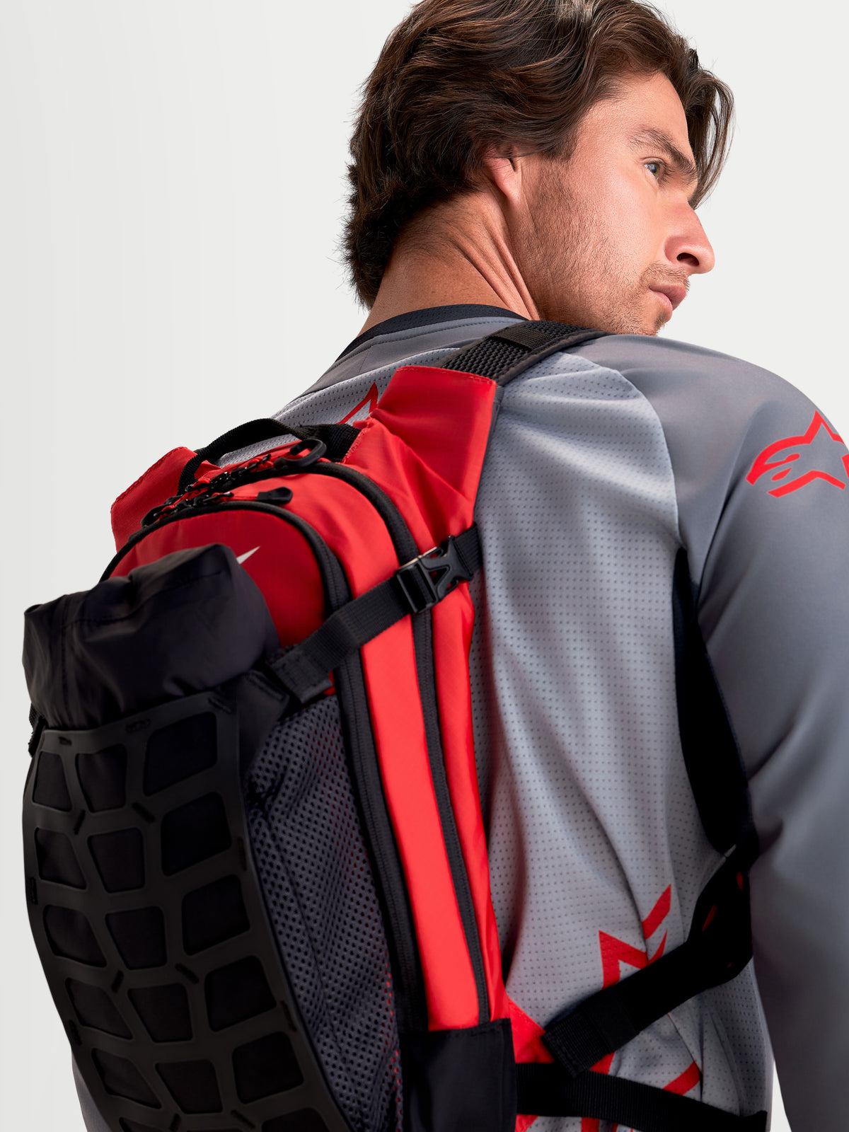 Alpinestars Mochila AT-9, mochila de motocross preta e vermelha, vista do perfil traseiro usada por um modelo a exibir construção têxtil leve e alças de ombro acolchoadas