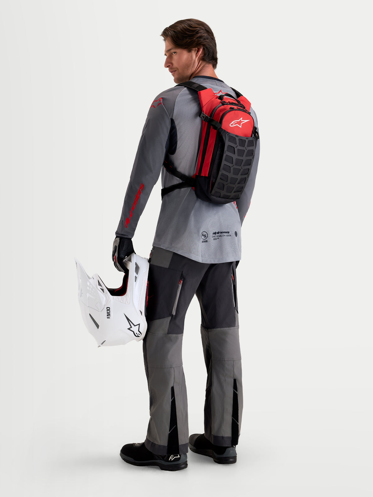 Alpinestars Mochila AT-9, mochila de motocross em preto e vermelho vivo, vista traseira num modelo a segurar um capacete de MX branco e equipamento a combinar, construção têxtil leve para utilização em pista e paddock