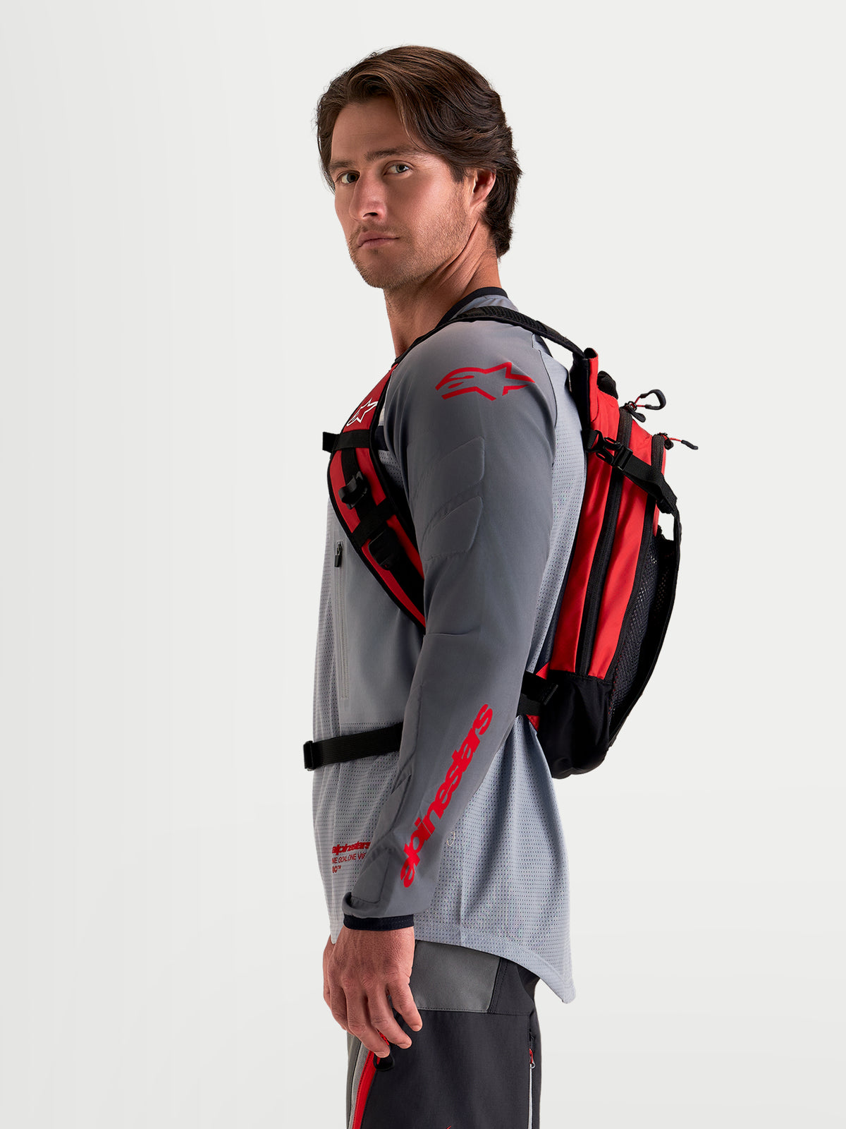 Alpinestars Mochila AT-9, mochila de motocross para homem, preta e vermelho vivo, vista de perfil lateral num modelo a usar equipamento MX a combinar, com construção leve em tecido e sistema de arnês ergonómico