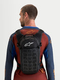 Alpinestars Mochila AT-9, mochila de motocross preta com logótipo branco, design compacto e ergonómico, vista traseira num modelo