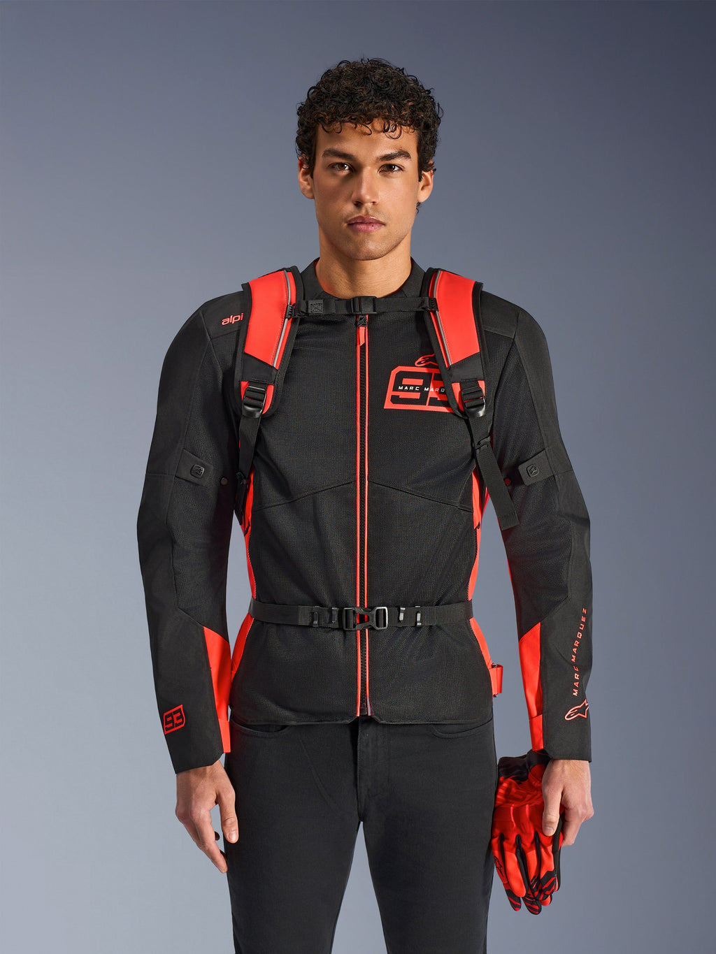 Homem a usar a mochila Alpinestars MM93 AMP3, em vermelho fluo e preto, com alças de ombro ajustáveis e correia de peito com fivela, combinada com casaco de motociclismo e luvas MM93 a condizer, equipamento técnico da coleção Marc Marquez
