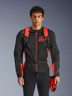Homem a usar a mochila Alpinestars MM93 AMP3, em vermelho fluo e preto, com alças de ombro ajustáveis e correia de peito com fivela, combinada com casaco de motociclismo e luvas MM93 a condizer, equipamento técnico da coleção Marc Marquez