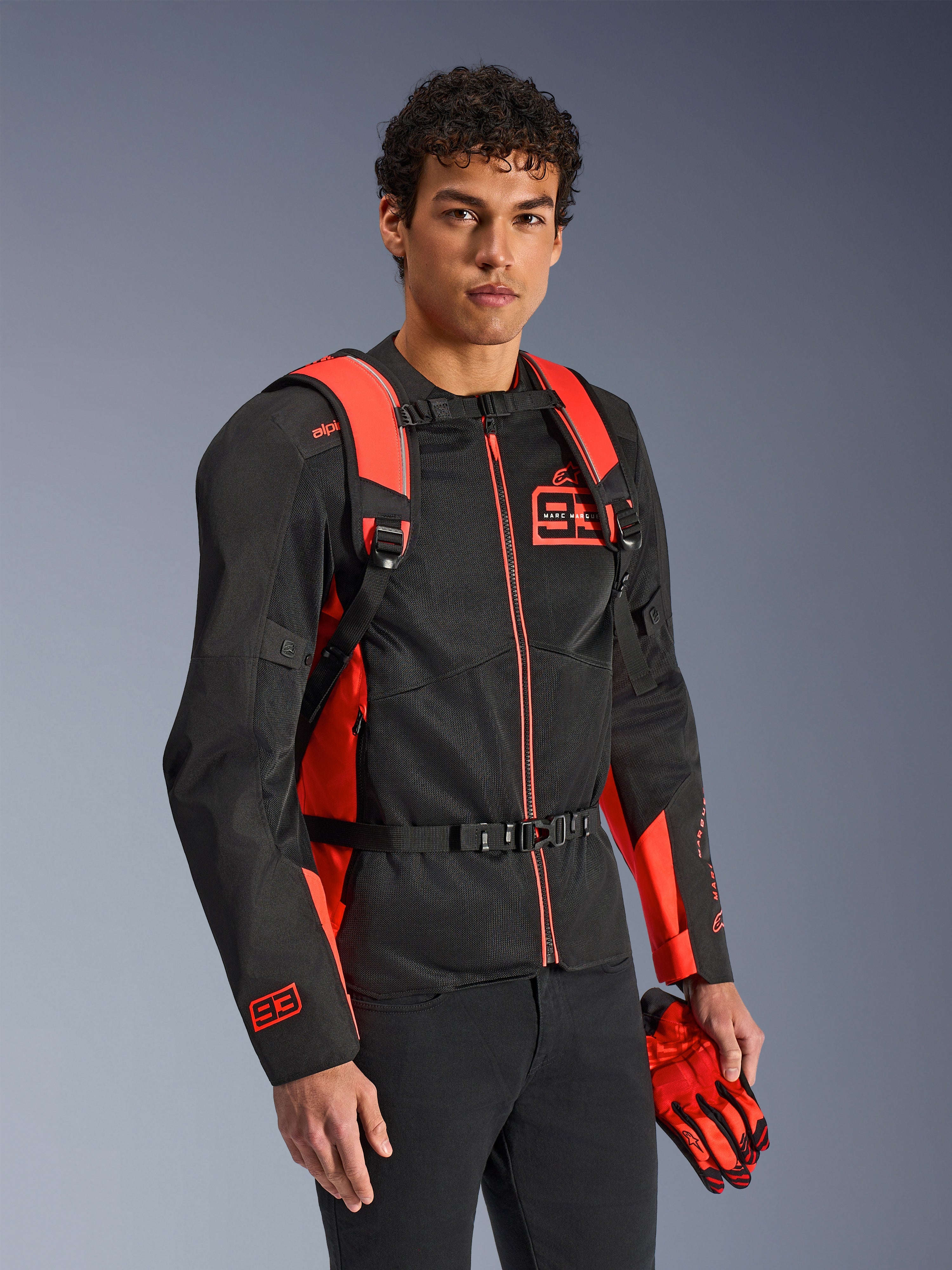 Mochila Alpinestars MM93 AMP3, vermelho fluo e preto, usada por um modelo com casaco e luvas de motociclismo MM93 a condizer em preto e vermelho, vista frontal destacando as alças ajustáveis e a fivela no peito, equipamento técnico inspirado em competição