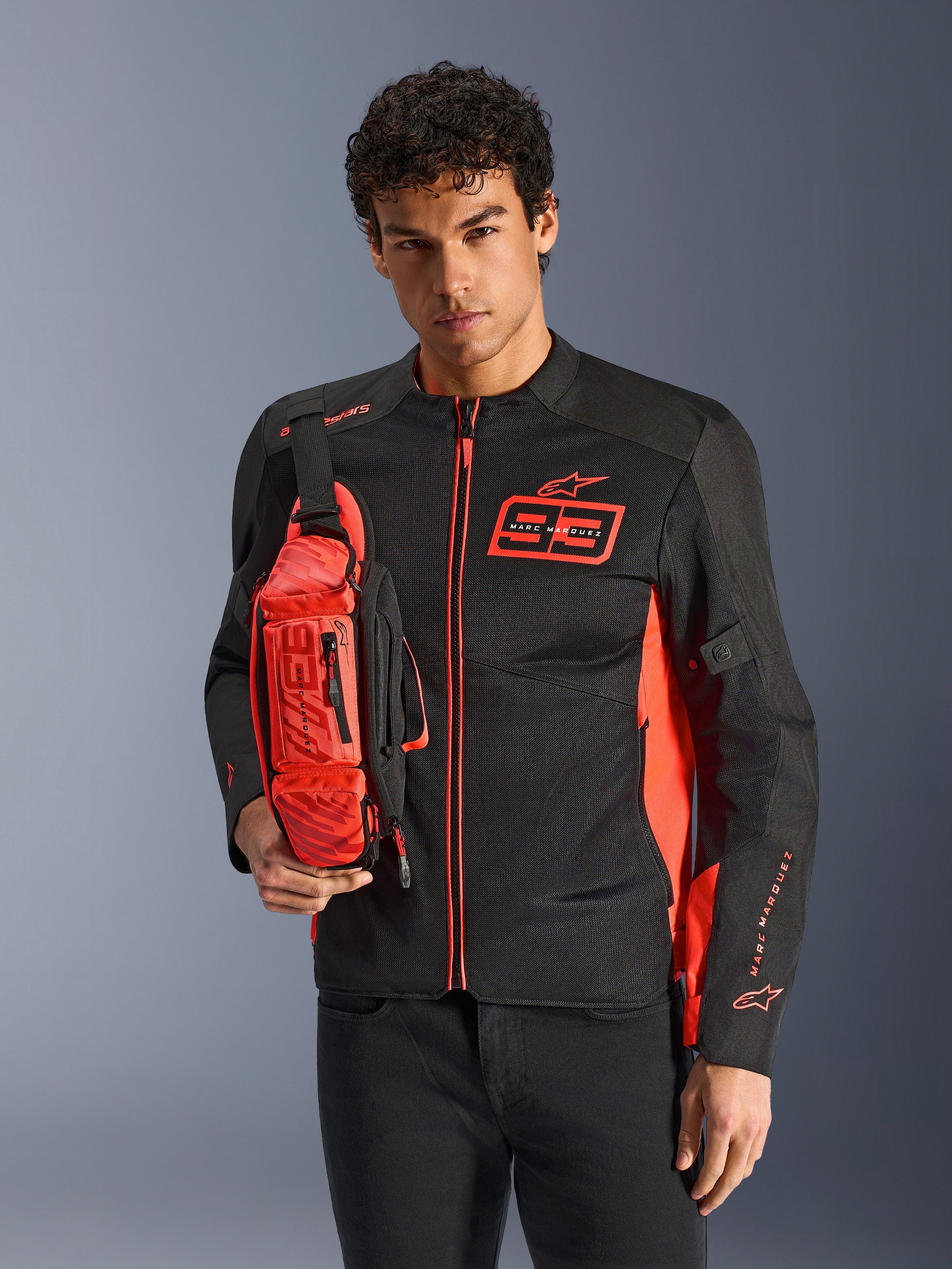 Homem a usar a Alpinestars MM93 Waist Bag, em vermelho fluo e preto, usada como mala tiracolo sobre um blusão de motociclismo preto e vermelho a condizer, com o logótipo Marc Marquez 93 e design desportivo de competição