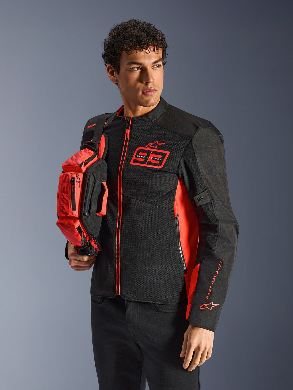 Alpinestars MM93 Waist Bag, bolsa de cintura em Vermelho Fluo e Preto, segurada por um modelo a usar um casaco de motociclismo preto e vermelho a condizer, com vários compartimentos com fecho e marca Marc Marquez, equipamento prático para condução desportiva