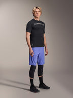 Modelo masculino a usar equipamento de mountain bike Alpinestars, com a camisola preta A-Aria Switch SS e os calções A-Aria Lite em Baja Blue, combinados com joelheiras pretas, meias pretas e sapatos de mountain bike pretos, numa fotografia de estúdio de corpo inteiro.