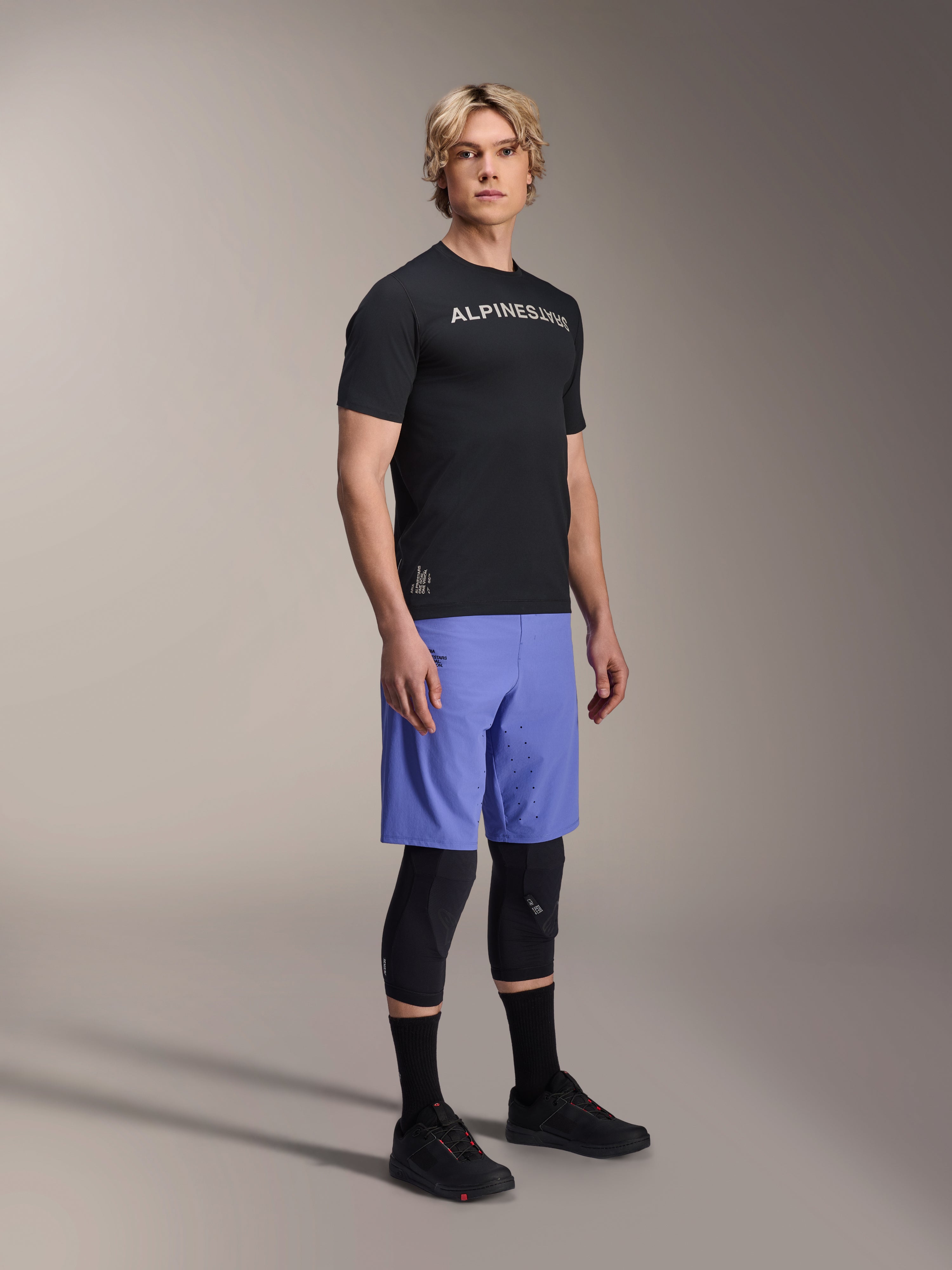 Modelo masculino a usar a camisola de manga curta Alpinestars A-Aria Switch em preto com logótipo branco no peito, combinada com calções de BTT azul-claro, joelheiras pretas e sapatos de ciclismo pretos, fotografia de corpo inteiro em estúdio para equipamento de BTT de alta performance
