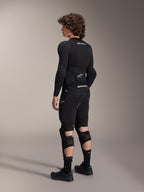 Modelo a usar os Protetores de Joelho e Canela Alpinestars A-Impact Plasma Pro, vistos de costas, em preto e branco, com sistema de fecho de tripla tira e painéis em malha ventilada, usados com equipamento completo de proteção de BTT preto, incluindo camisola interior de manga comprida e calções acolchoados.