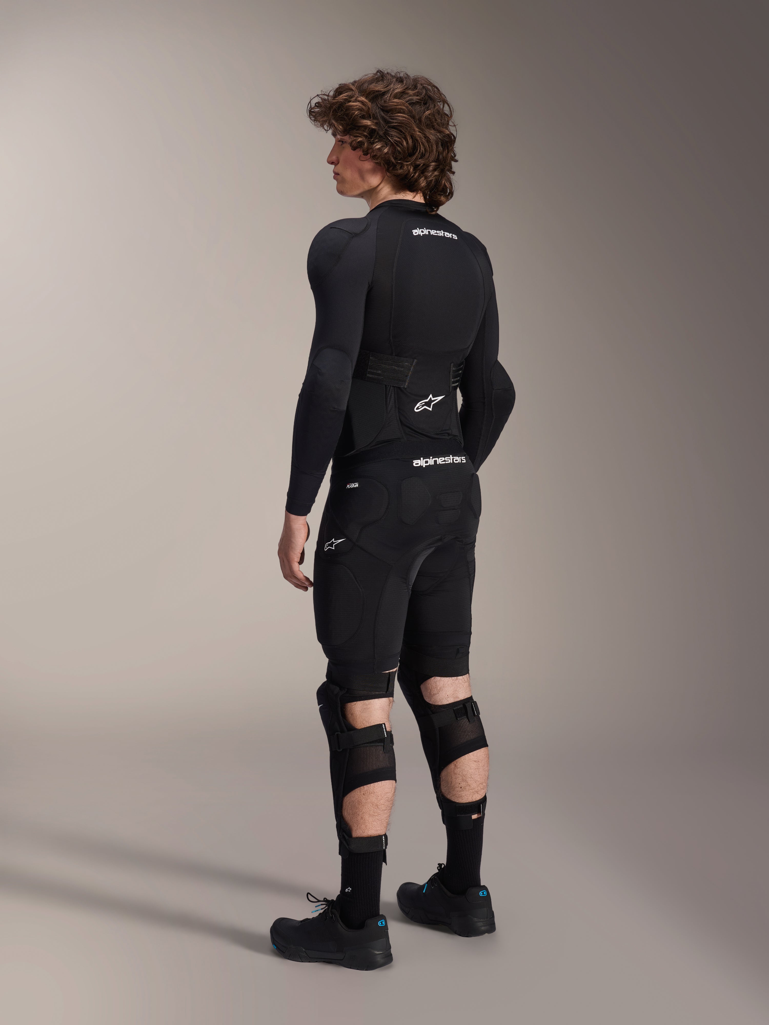 Modelo a usar os Protetores de Joelho e Canela Alpinestars A-Impact Plasma Pro, vistos de costas, em preto e branco, com sistema de fecho de tripla tira e painéis em malha ventilada, usados com equipamento completo de proteção de BTT preto, incluindo camisola interior de manga comprida e calções acolchoados.