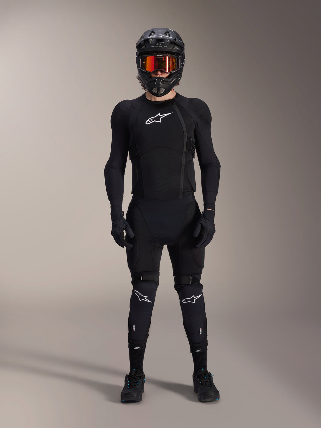Ciclista de mountain bike com equipamento de proteção completo, Casaco de Proteção Alpinestars A-Impact Plasma em preto e branco, com logótipo branco no peito e tecido de malha, usado com capacete preto, óculos de lentes avermelhadas e joelheiras, conjunto de armadura de BTT de alta performance coordenado.