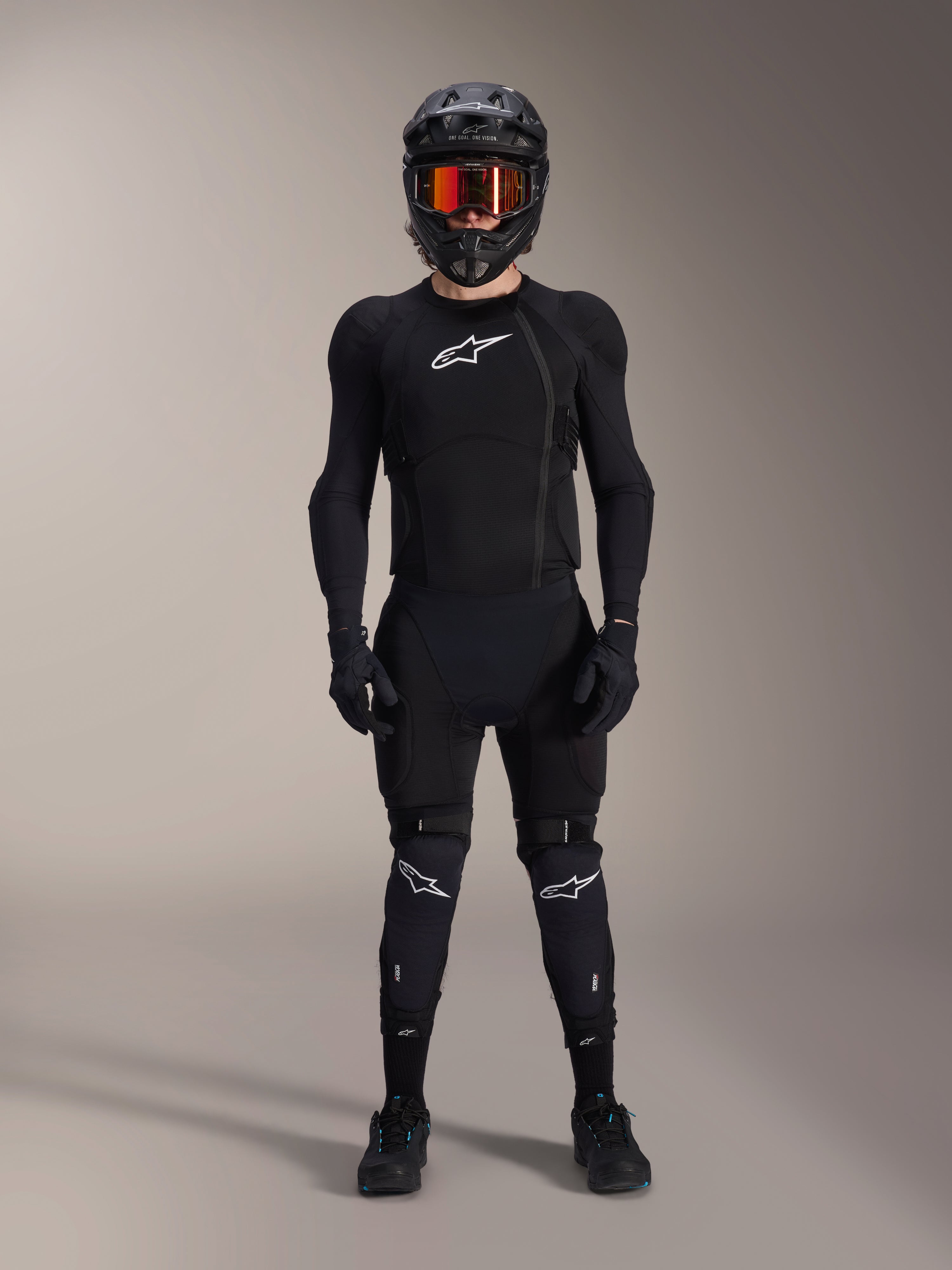 Ciclista de mountain bike com equipamento de proteção completo, Casaco de Proteção Alpinestars A-Impact Plasma em preto e branco, com logótipo branco no peito e tecido de malha, usado com capacete preto, óculos de lentes avermelhadas e joelheiras, conjunto de armadura de BTT de alta performance coordenado.