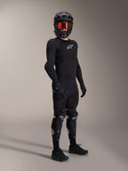 Protetores de Joelho e Canela Alpinestars A-Impact Plasma Pro, pretos e brancos, usados por um ciclista com equipamento de proteção completo para mountain bike, incluindo camisola blindada preta de manga comprida, luvas pretas e capacete integral com óculos cor de laranja