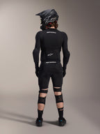 Protetores de Joelho e Canela Alpinestars A-Impact Plasma Pro, Pretos, vista traseira de um ciclista com equipamento de proteção completo de mountain bike, incluindo capacete integral e proteção de corpo, com sistema de fecho de três tiras e painéis de malha ventilada para um ajuste seguro e ventilação