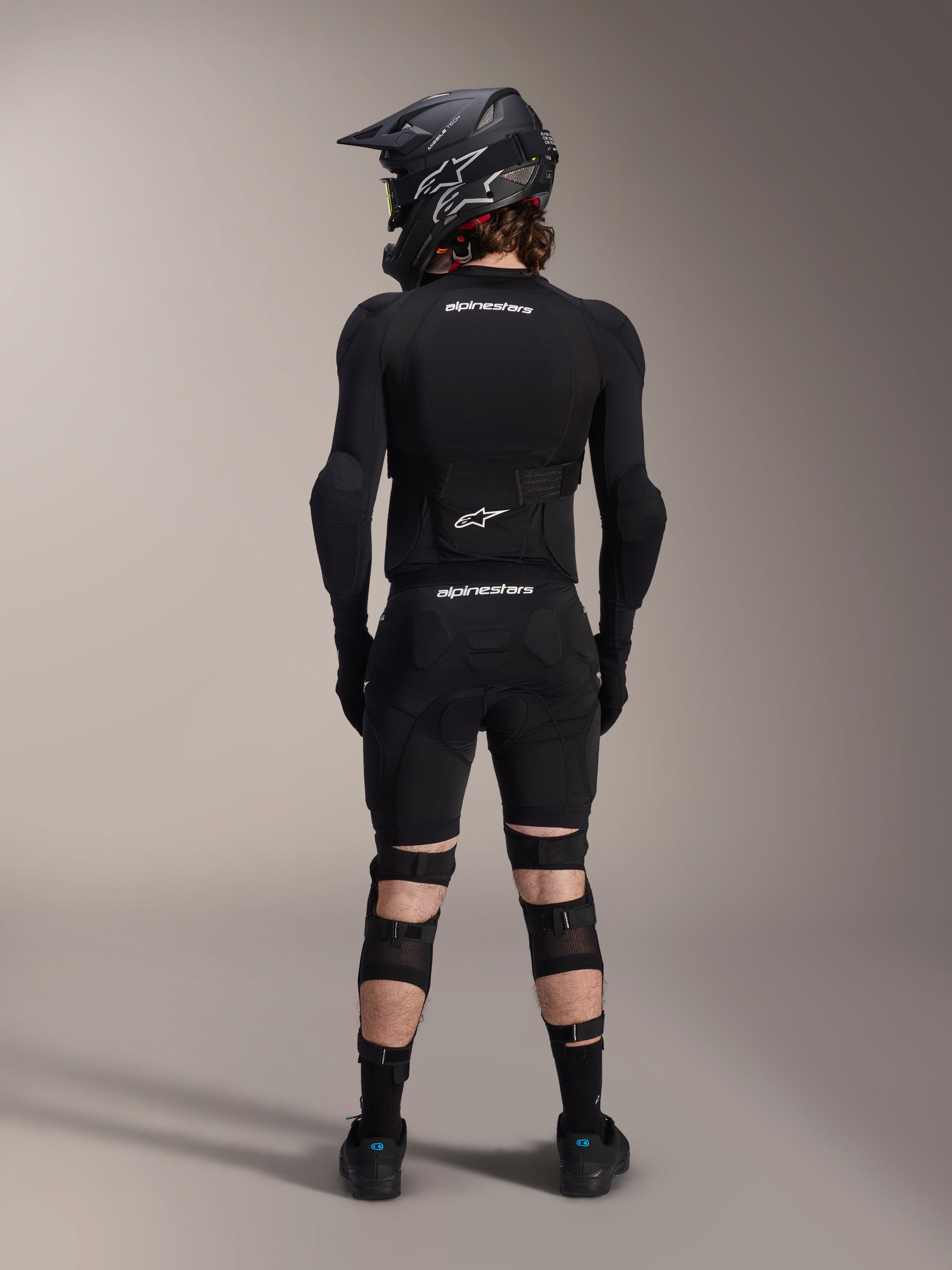 Protetores de Joelho e Canela Alpinestars A-Impact Plasma Pro, Pretos, vista traseira de um ciclista com equipamento de proteção completo de mountain bike, incluindo capacete integral e proteção de corpo, com sistema de fecho de três tiras e painéis de malha ventilada para um ajuste seguro e ventilação