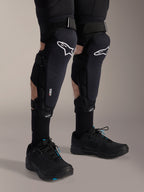 Protetores de joelho e canela Alpinestars A-Impact Plasma Pro, pretos e brancos, com o logótipo Alpinestars branco e sistema de fecho de três tiras, usados com meias pretas e sapatilhas desportivas pretas, proteção durável para ciclismo fora de estrada
