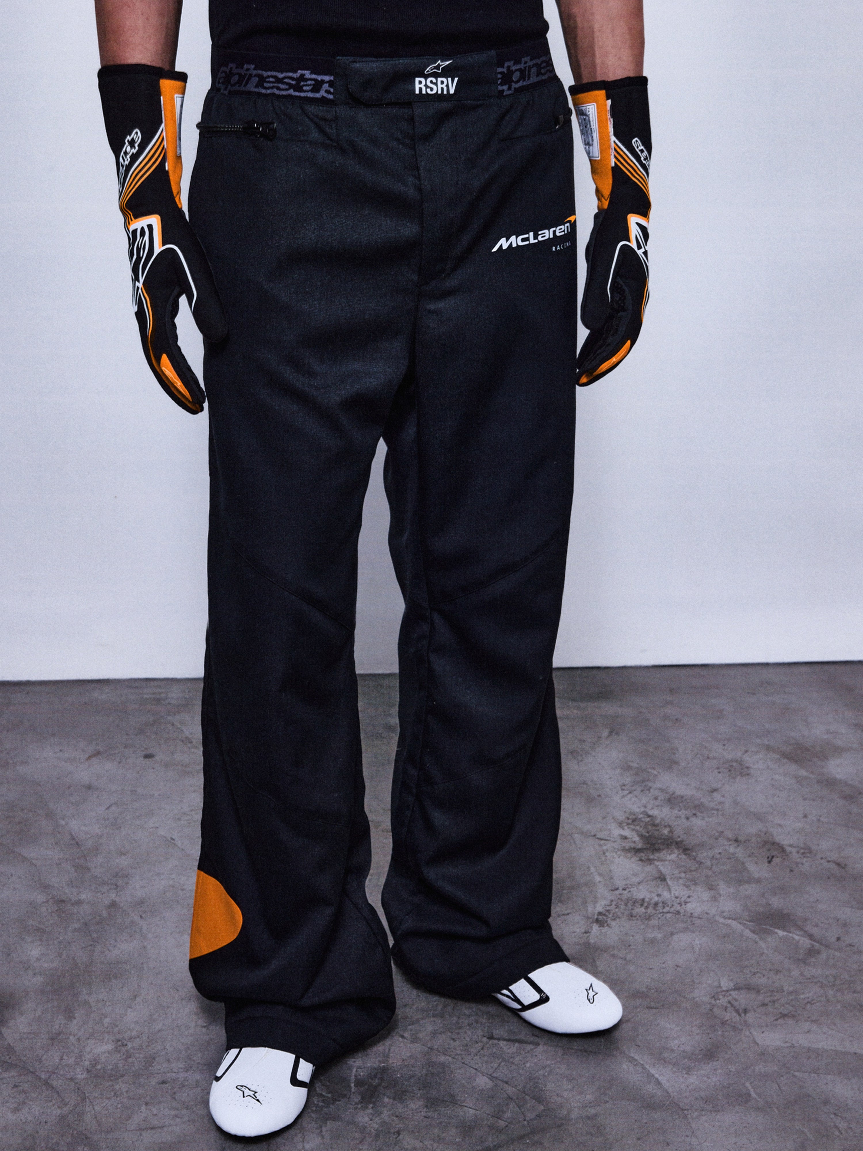 RSRV Arena Suit Pants, Black Racing Pants with McLaren and Alpinestars Logos, Orange Side Patches, Premium Motorsport Apparel, Worn with Black and Orange Gloves and White Racing Shoes