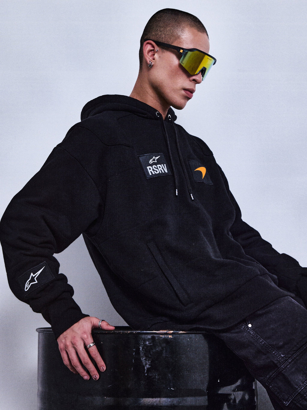 Alpinestars RSRV Rival Hoodie, moletom preto unissexo com logótipo RSRV e patches, estilo streetwear premium, pessoa com óculos de sol refletores e anéis sentada em barril metálico
