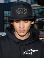 Boné Trucker Alpinestars Contender, boné casual preto com malha de poliéster na parte traseira, copa estruturada com remendo bordado do logótipo Astars, usado por um homem com hoodie preto Alpinestars, streetwear casual inspirado no desporto motorizado.