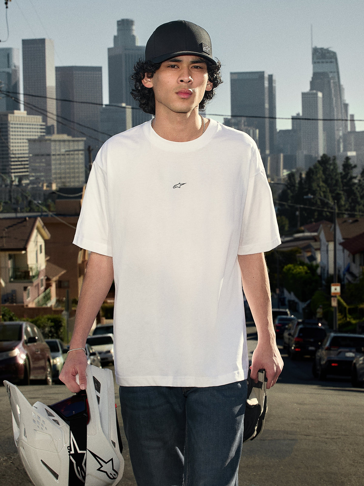 T-shirt Alpinestars Focused Oversized CSF, t-shirt de manga curta, branca, com um pequeno logótipo Astars preto no peito, fotografia de estilo de vida de uma pessoa a usar a t-shirt e a segurar um capacete de motocross com o horizonte da cidade ao fundo, streetwear casual de desportos motorizados