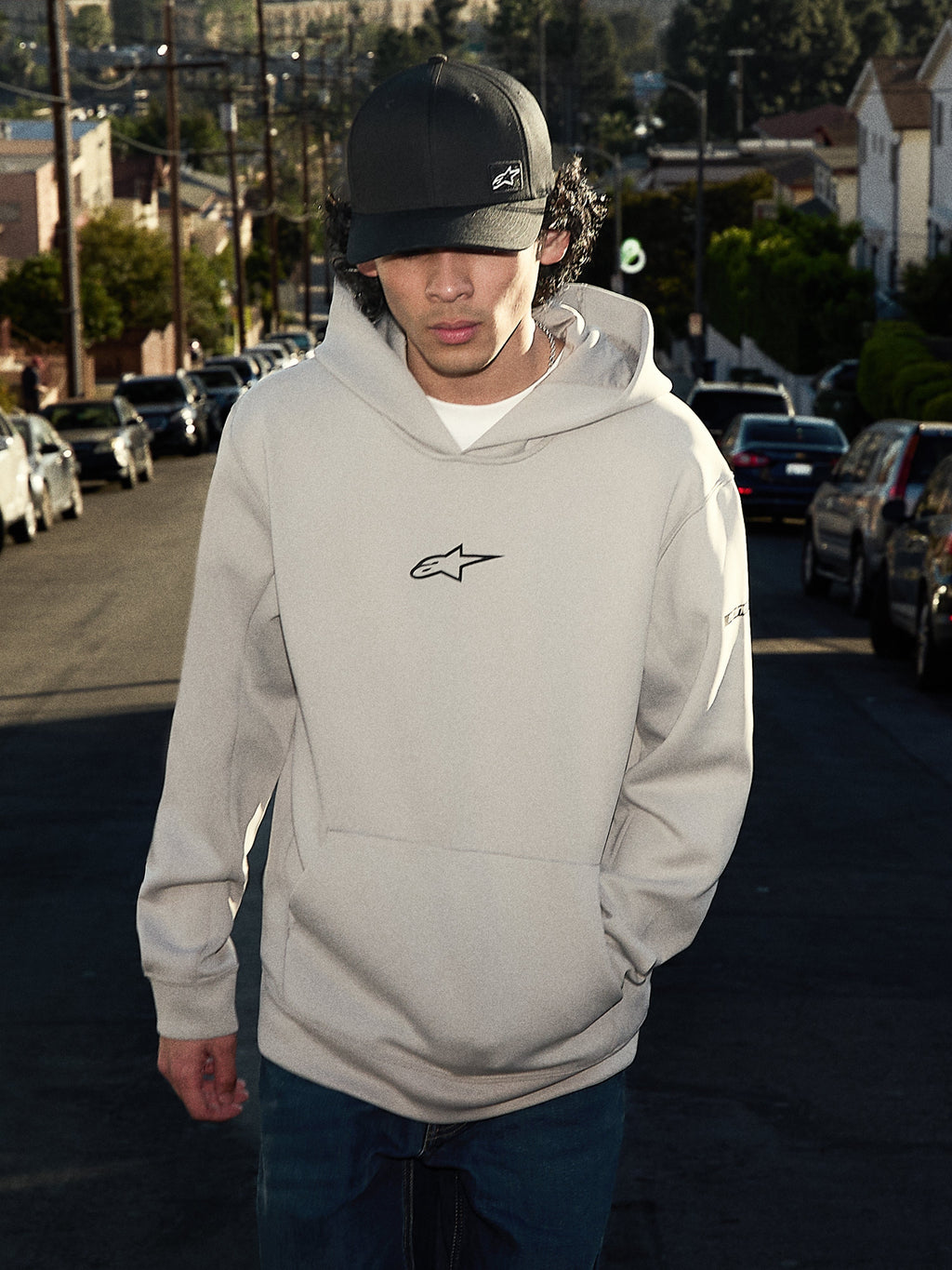 Alpinestars Insight Hoodie, sweatshirt de estilo casual, cinzento Cement com logótipo Astars preto no peito, modelo pullover com bolso canguru, usada com um boné preto numa rua residencial, design moderno inspirado no desporto motorizado