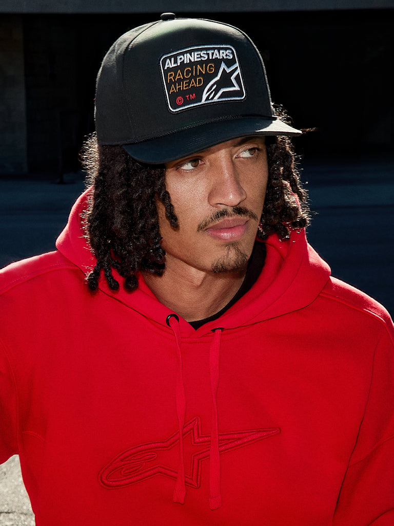 Homem a usar o boné Alpinestars Intuitive Snapback em preto, com copa estruturada e patch bordado com o logótipo 'Racing Ahead', combinado com um hoodie vermelho vibrante, estilo casual lifestyle streetwear
