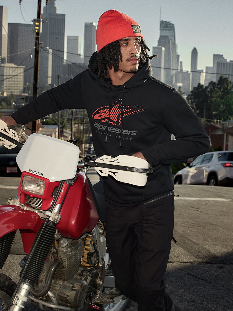 Alpinestars Pulse Hoodie, camisola com capuz preta com logótipo Astars Combo vermelho e cinzento, usada por um homem com um gorro cor de laranja ao lado de uma mota Honda vermelha num cenário urbano, streetwear casual de desportos motorizados