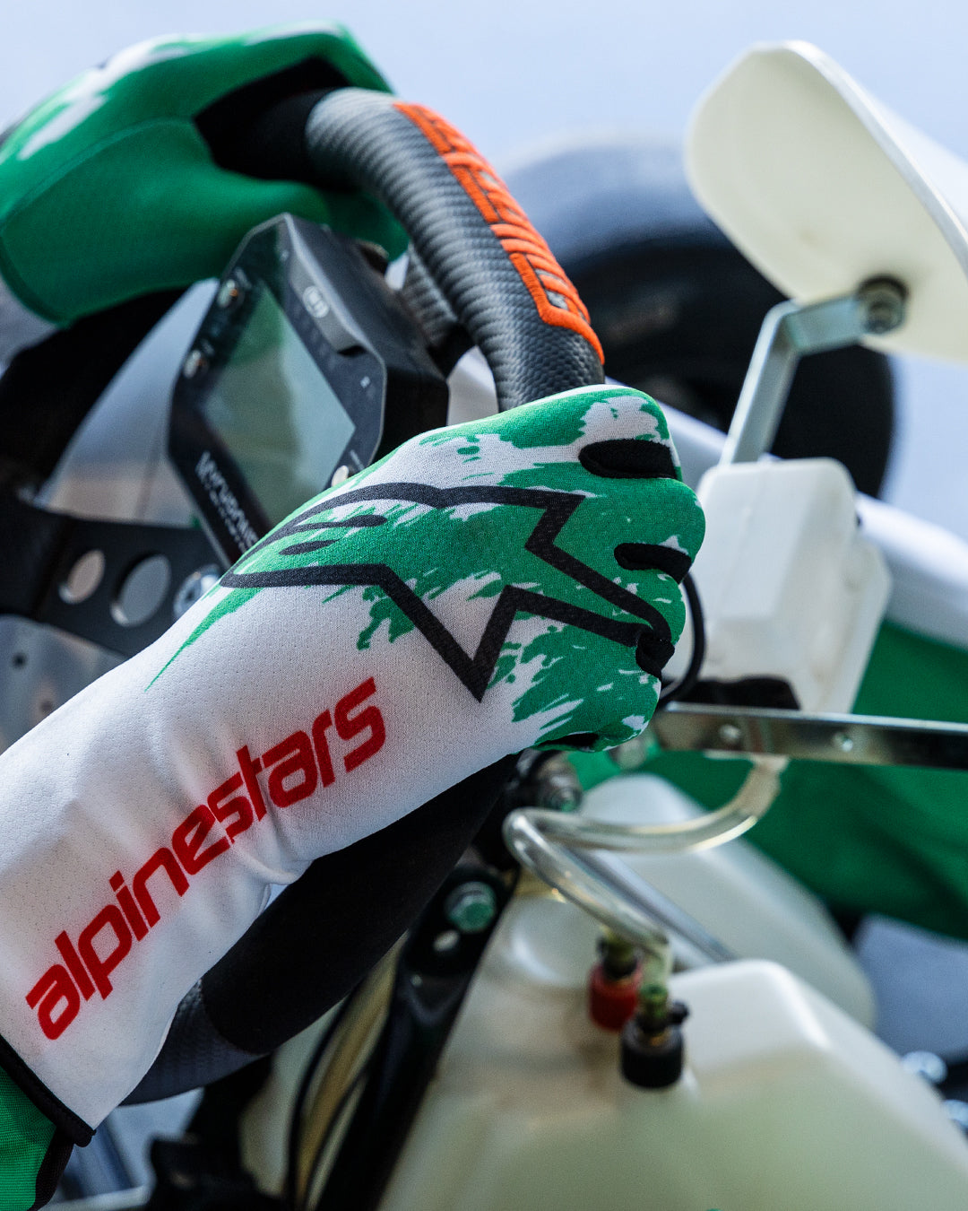 Luvas Alpinestars Tech-1 K V3 em branco, verde e vermelho, mãos do piloto a segurar um volante de karting, com um design de salpicos sublimados a verde, logótipo da estrela a preto e texto Alpinestars a vermelho no punho, equipamento profissional de karting.