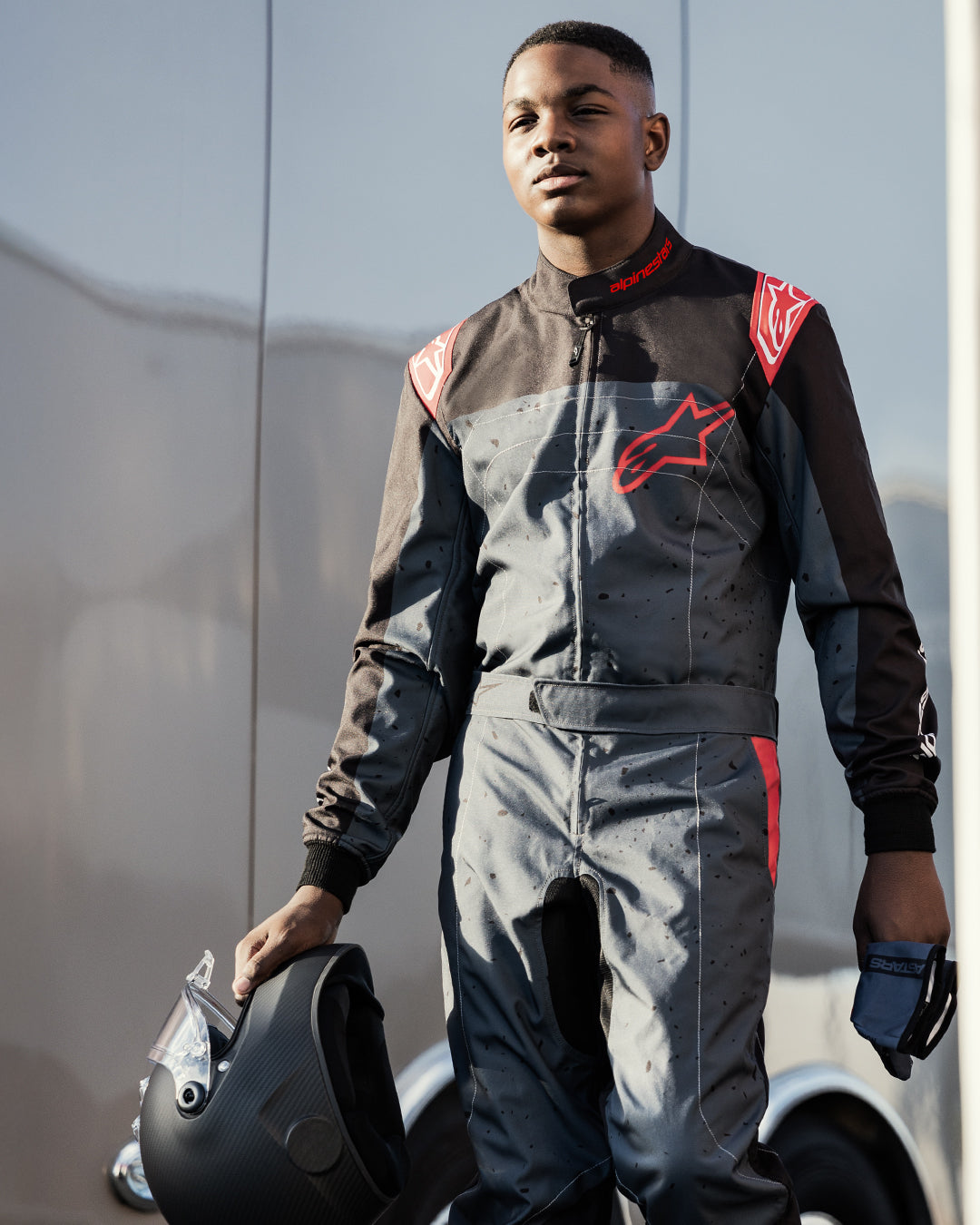 Piloto de kart a usar o fato Alpinestars KMX-9 V3 Graphic 6 em preto antracite e vermelho, a segurar um capacete preto mate e luvas, com logótipos Alpinestars a vermelho em destaque e um design gráfico com efeito salpicado, num cenário de desporto motorizado ao ar livre.