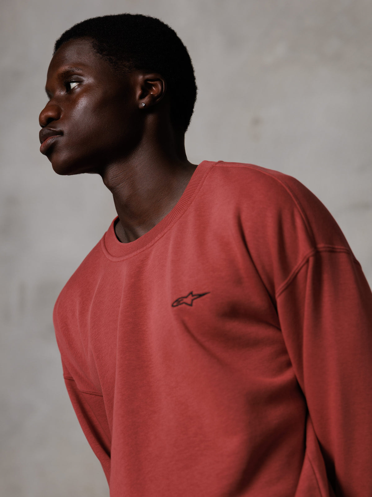 Alpinestars Essentials Ageless Crew, sweatshirt de gola redonda em vermelho tijolo, vestida por um modelo masculino de perfil, pequeno logótipo de estrela preto no peito, corte descontraído, fleece premium, streetwear casual