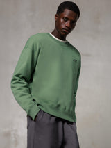 Alpinestars Essentials Ageless Crew, sweatshirt de gola redonda, verde cacto, com um pequeno logótipo preto da Alpinestars no peito, usada por um modelo com calças cinzentas, estilo streetwear casual