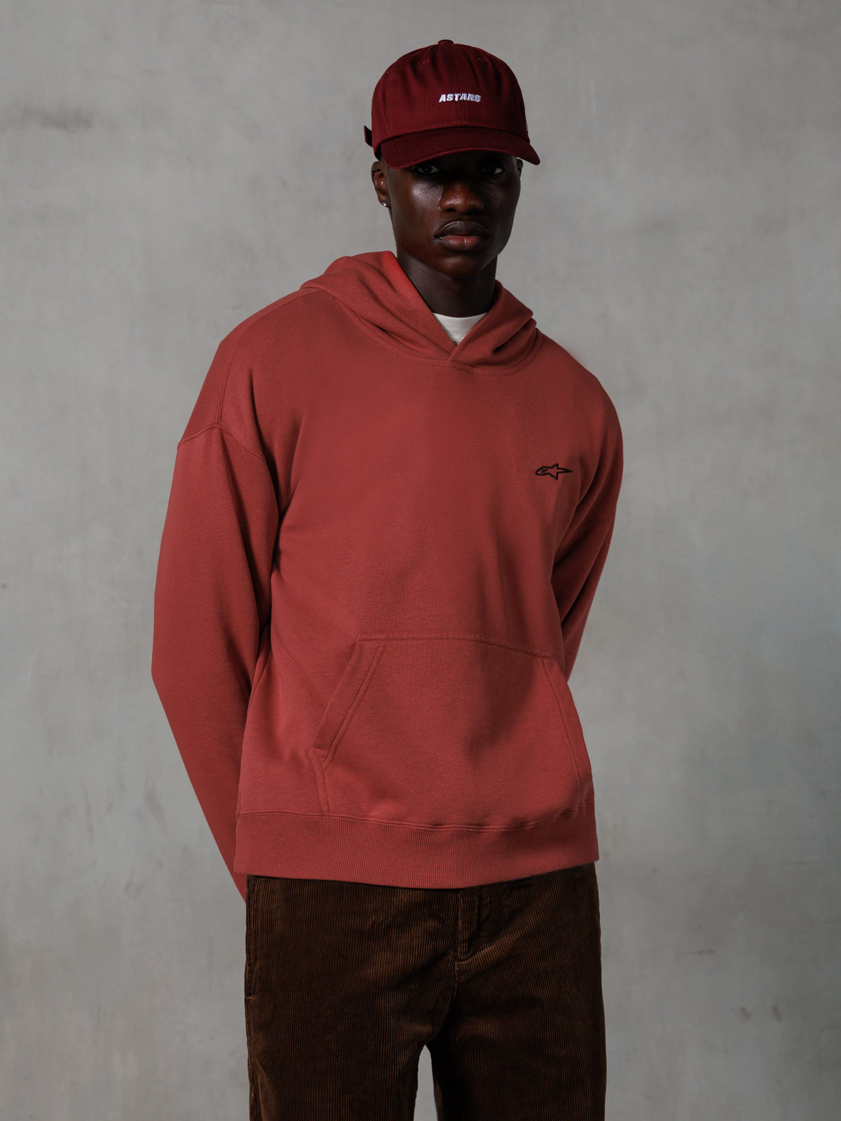 Hoodie Alpinestars Essentials Ageless, streetwear casual, cor vermelho tijolo, pequeno logótipo preto no peito, bolso canguru frontal, usado com um boné ASTARS vermelho a combinar e calças de bombazina castanhas, design minimalista e clean