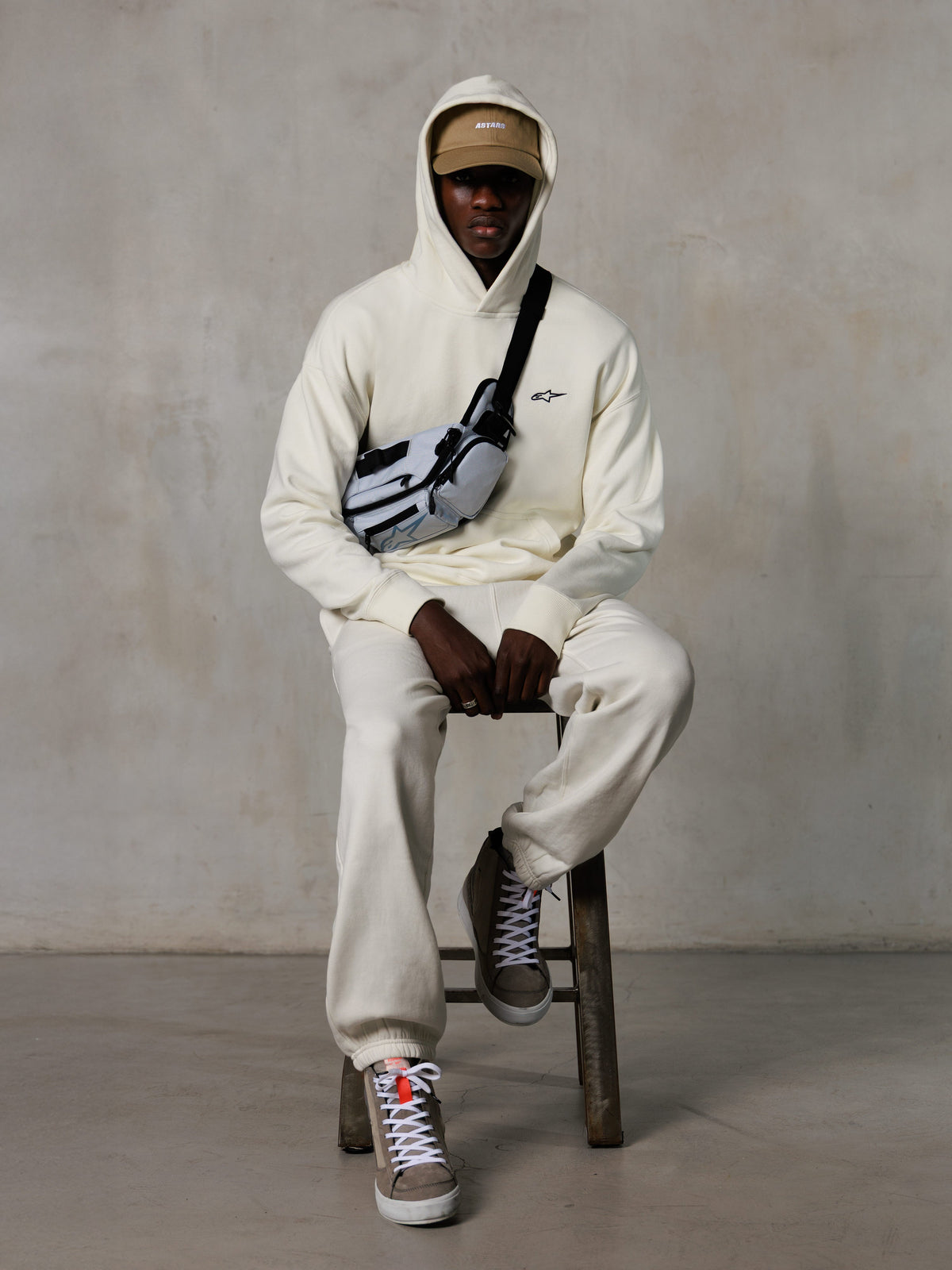 Alpinestars Essentials Ageless Hoodie, Off-White, usado por um modelo sentado num banco com calças de fleece a condizer, boné ASTARS bege e bolsa a tiracolo, streetwear casual inspirado no desporto motorizado